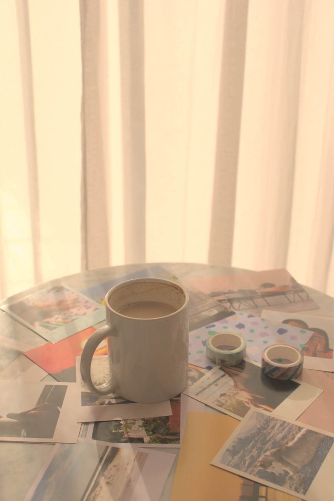White coffee cup, washi tapes, and printed custom photos, over a marble coffee table. The afternoon light filtering through the curtains in the background creates light and shadows over the coffee table.
