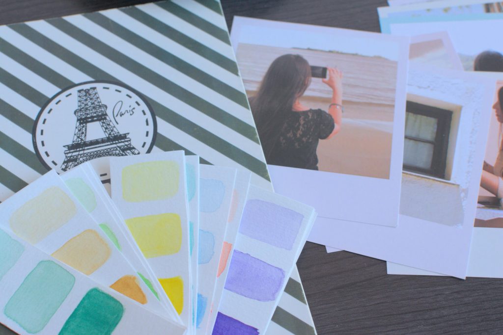 Handmade watercolor palette, a cute notebook, and printed custom photos, over a wooden table.