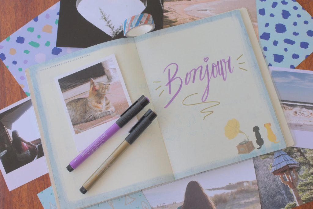 Flat lay of a cute notebook with the word “Bonjour”, and two metallic markers in purple and gold that were used to write the word. There is also a printed photo of a cute cat looking to the side, and postcards and printed brand patterns around the notebook.