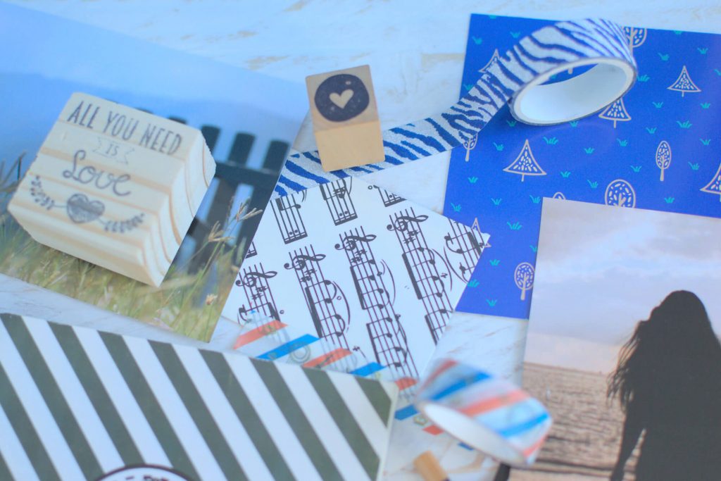 Close up a printed brand pattern with pine trees and grass. There are also washi tapes, a stamp with the words “All you need is love”, postcards, an envelope with a musical pattern, and a notebook. The background consists of white vintage wood.