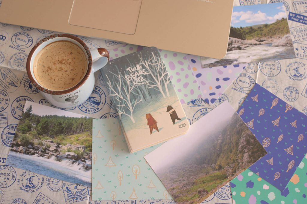 Flat lay of a light sky blue coffee cup, the corner of an open notebook in rose gold color, landscape postcards, printed brand patterns, and a cute notebook with bears. The background is a white wrapping paper with travel stamps in dark blue.